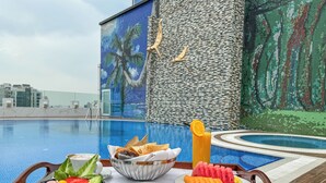 Bandeja de desayuno con panqueques, tostadas, fruta y jugo junto a una piscina con vista a la ciudad.