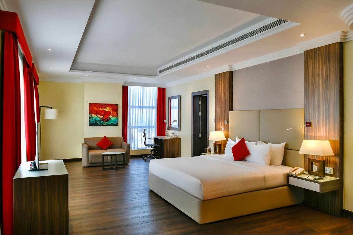 A hotel room with a large bed, a desk, a chair, and a sofa. There is a painting on the wall and a window with curtains.