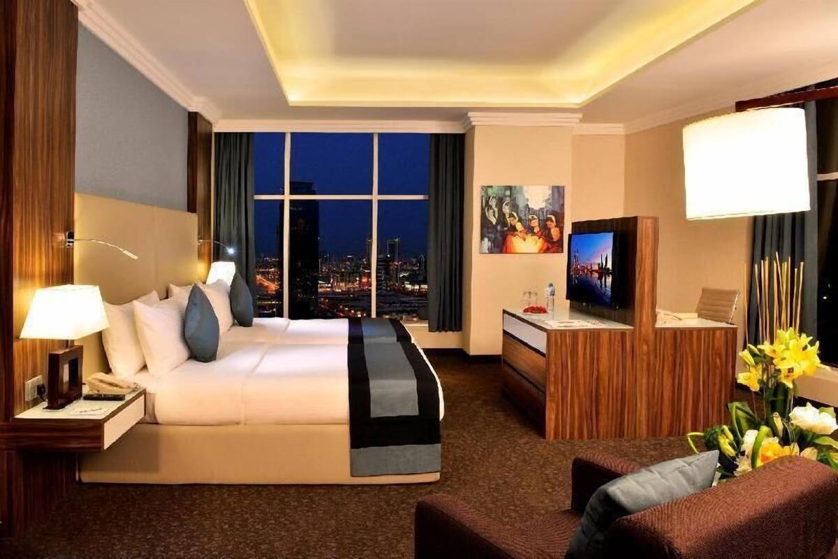 A hotel room with a bed, a desk, a TV, and a view of the city at night.