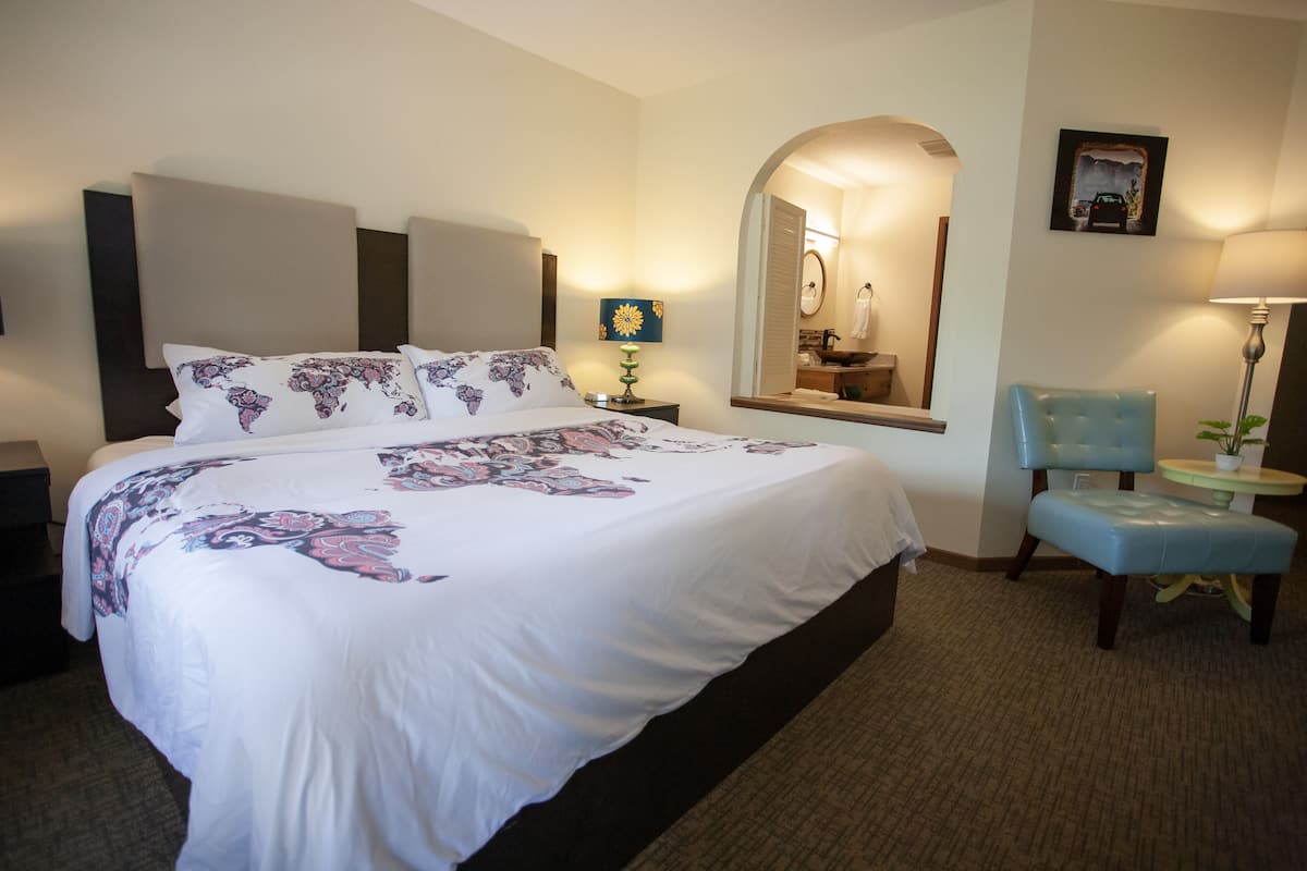 Habitación de hotel con una cama grande, una silla, una mesita, una lámpara, un espejo y un cuadro en la pared.