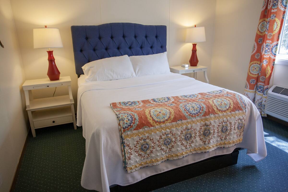 Una cama bien hecha con un edredón estampado, dos mesitas de noche con lámparas y una ventana con cortinas.