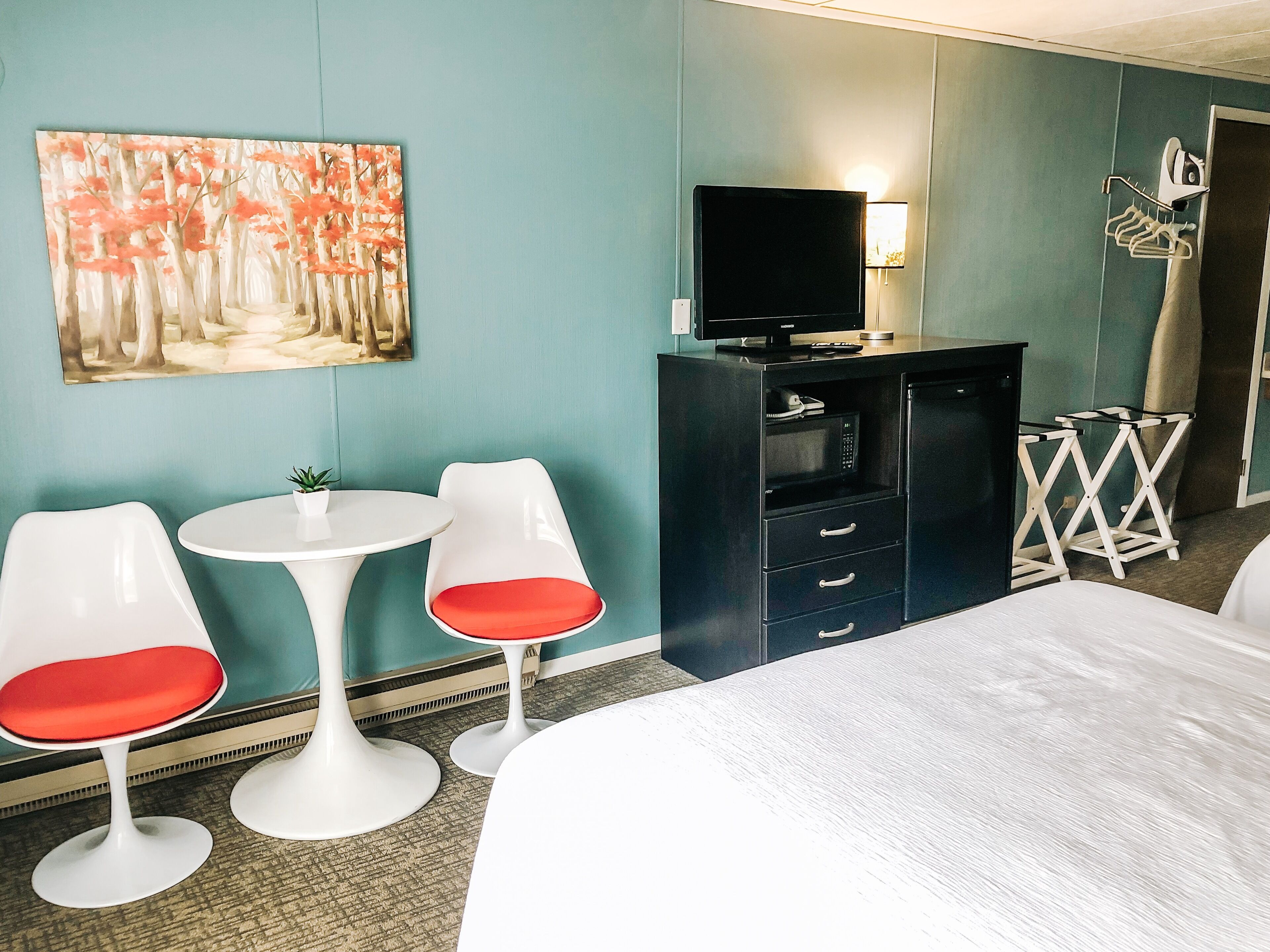 A hotel room with a bed, two white chairs with red cushions, a small table, a TV on a cabinet, and a painting on the wall.