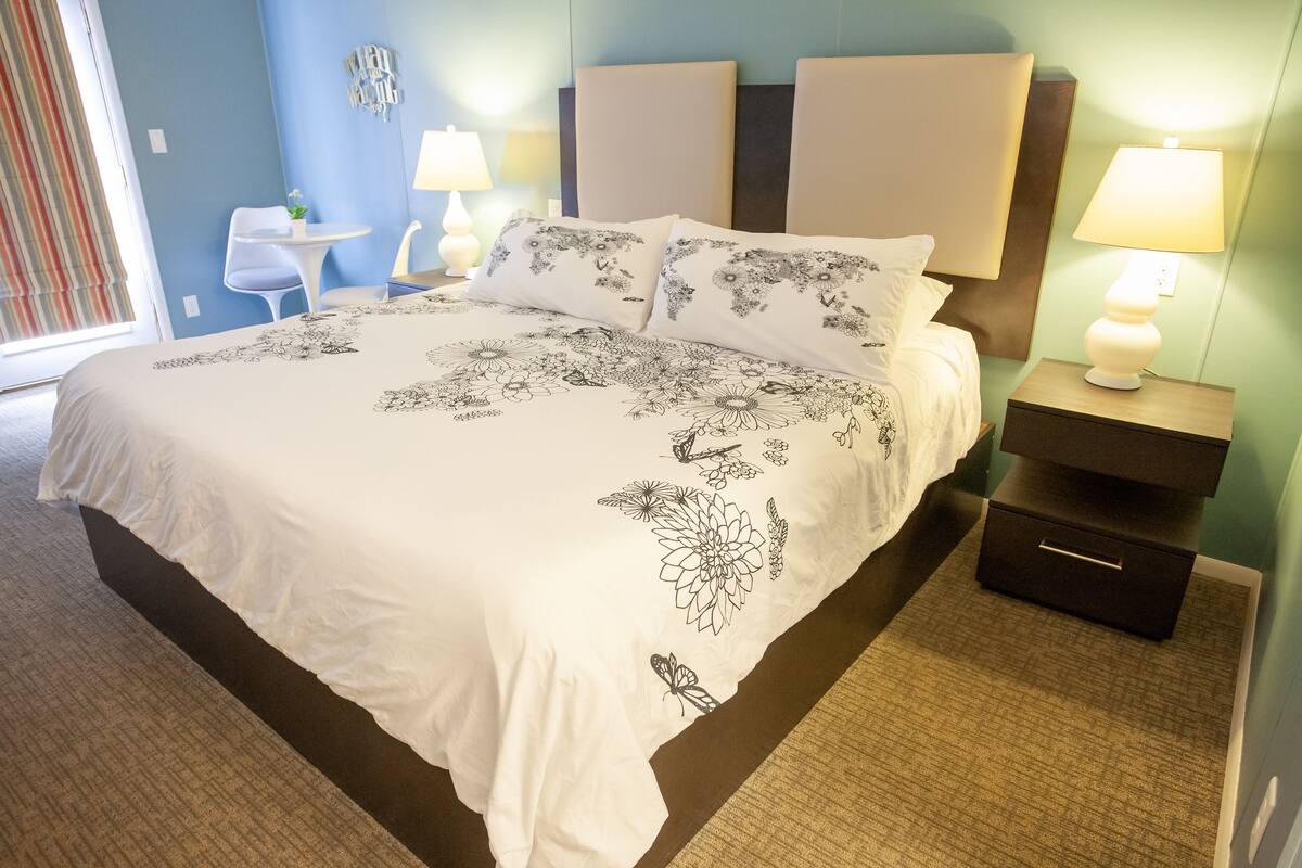 Una habitación de hotel con una cama grande, dos mesitas de noche con lámparas, una pequeña mesa blanca y una silla.