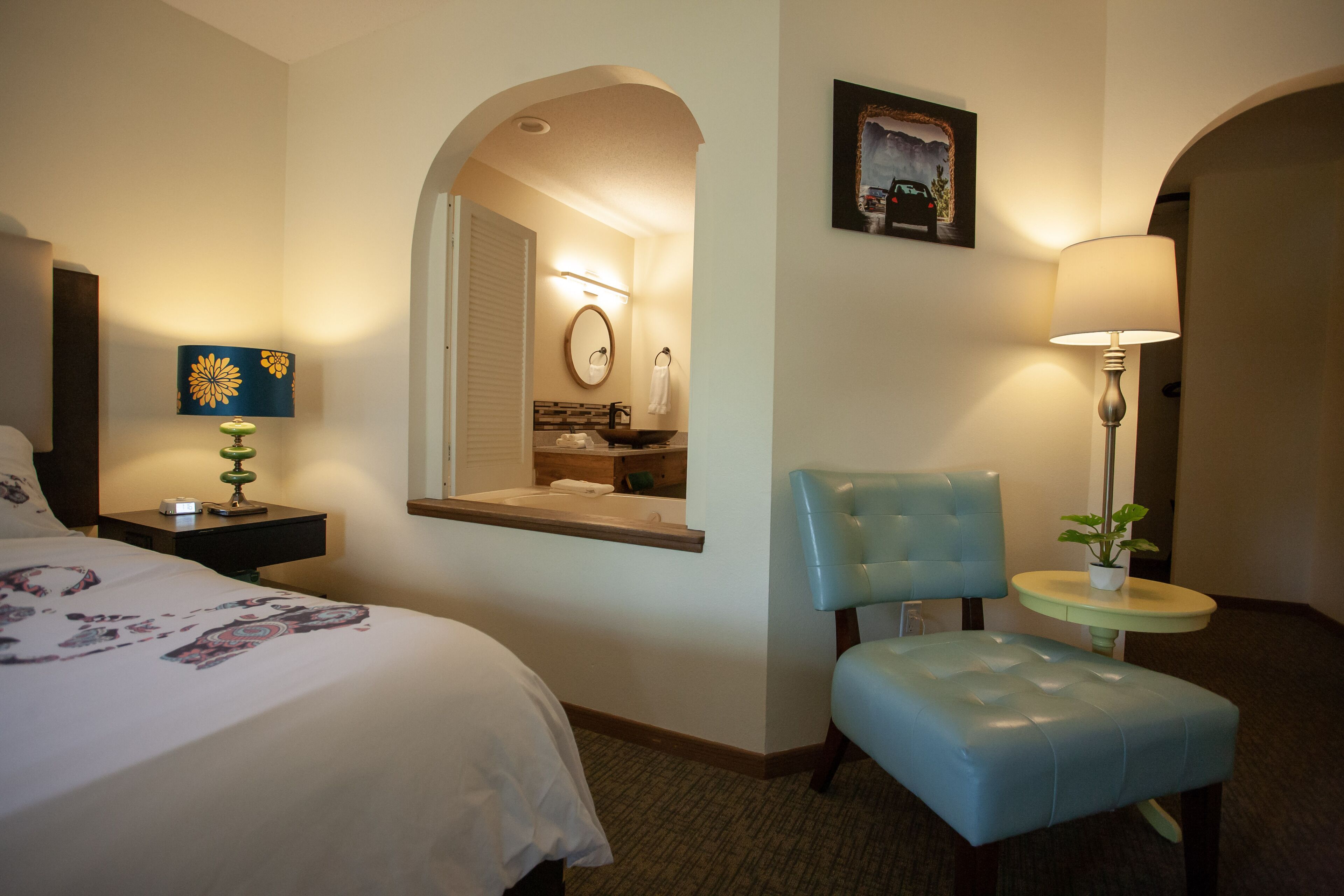 A hotel room with a bed, a small desk with a lamp, a chair, a round table with a plant, and a bathroom visible through an archway.