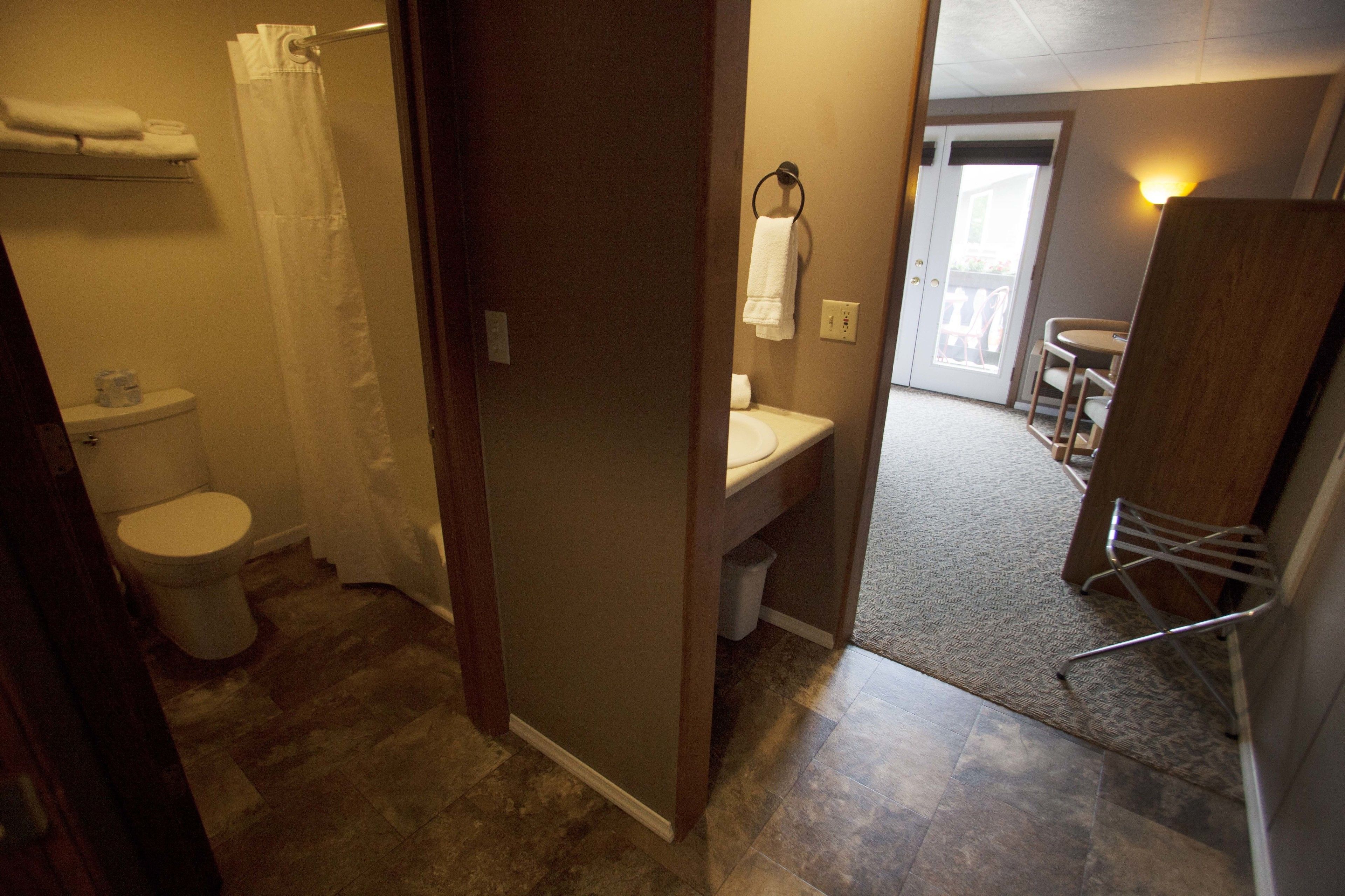 A bathroom with a toilet, sink, and shower curtain. There is a towel rack above the sink and a chair near the door.
