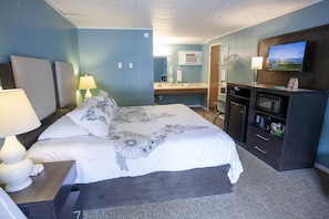 A hotel room with a large bed, a desk with a lamp, a TV, a microwave, and a bathroom visible in the background. - Bavarian Inn Black Hills (Custer)