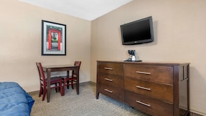 Ein Hotelzimmer mit einem Bett, einem Schreibtisch mit Stühlen, einem Schminktisch, einem Fernseher und einem gerahmten Bild an der Wand.