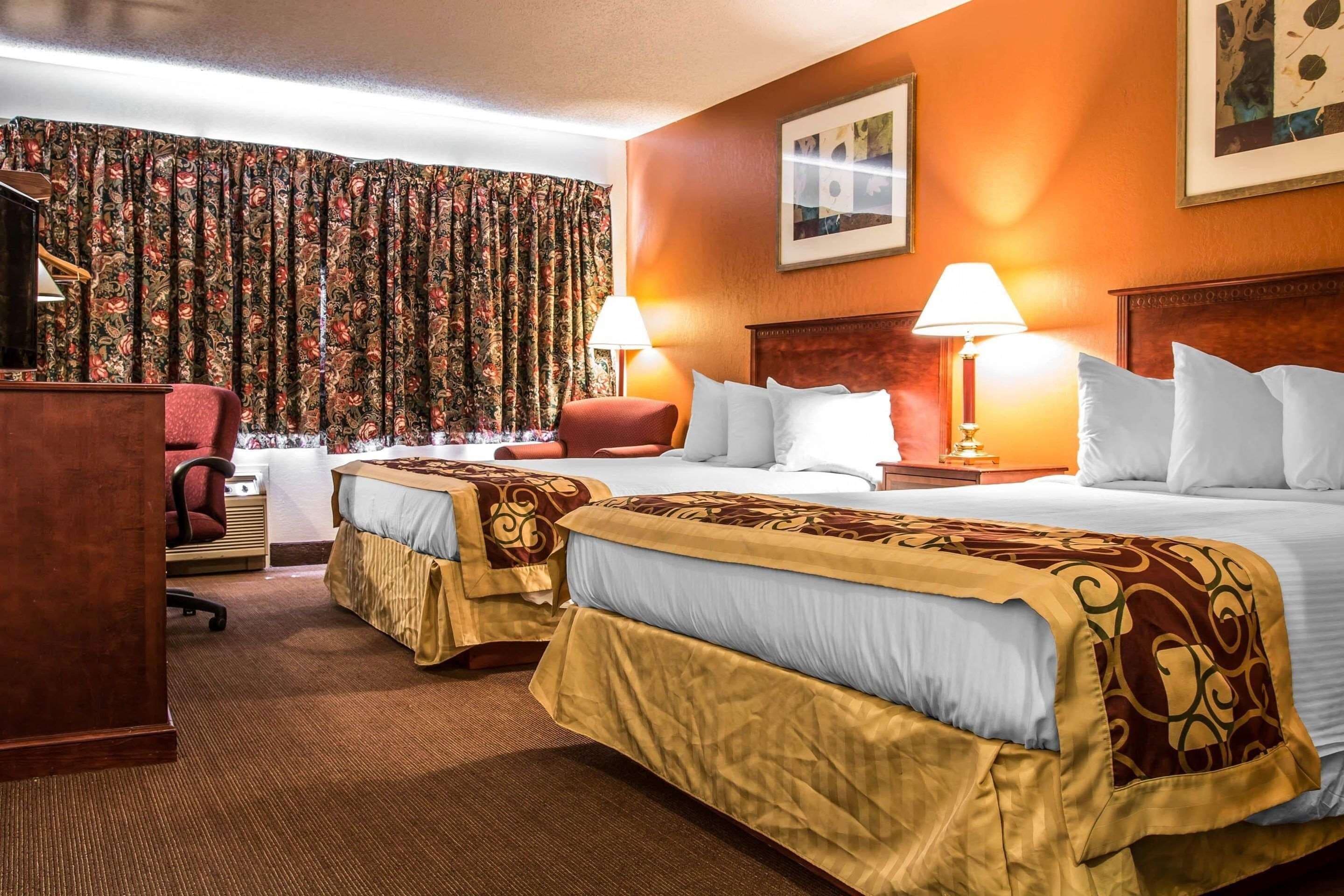 Une chambre d’hôtel avec deux lits, un bureau, une chaise, une lampe de bureau et une grande fenêtre avec des rideaux.