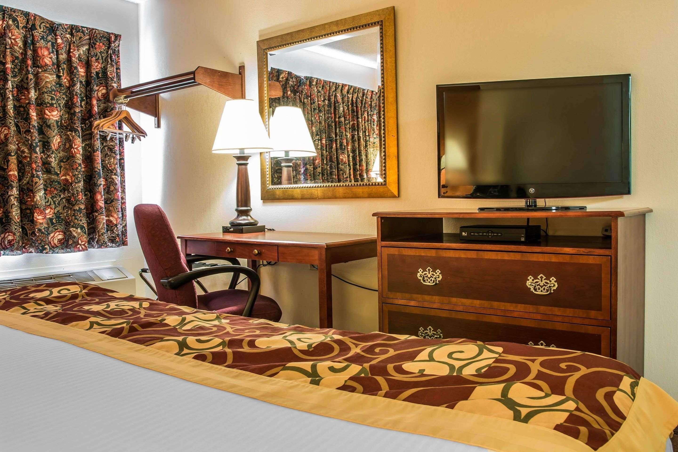 Quarto de hotel com cama, escrivaninha com cadeira, televisão, cómoda e espelho.