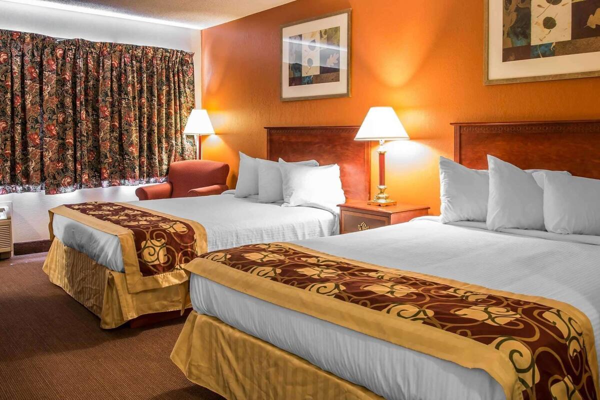 A hotel room with two beds, a red chair, a nightstand with a lamp, and a window with curtains.