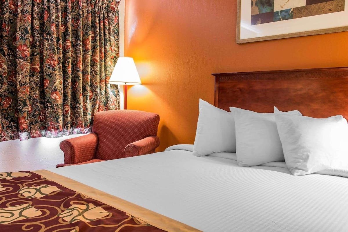 A hotel room with a bed, pillows, a chair, a lamp, and curtains.