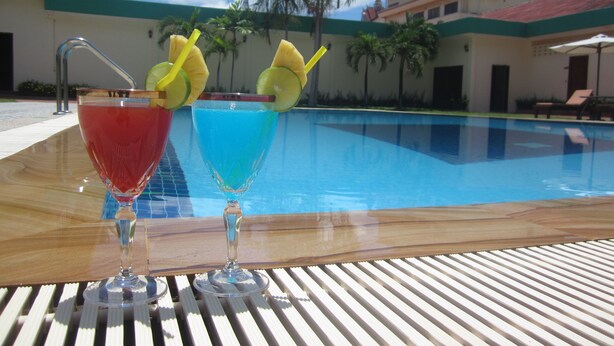 Dos cócteles sobre una mesa de madera junto a una piscina, con un cielo azul despejado y palmeras al fondo.
