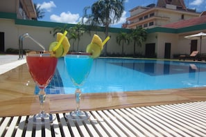 Two cocktails on a wooden table by a pool with a clear blue sky and palm trees in the background. - City Angkor Hotel (Siem Reap)