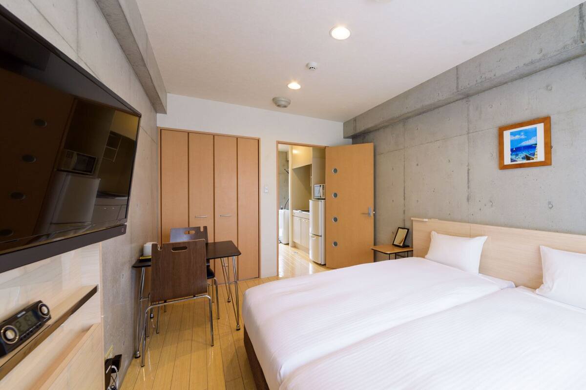 A hotel room with a bed, a desk, a chair, a TV, and a kitchenette.