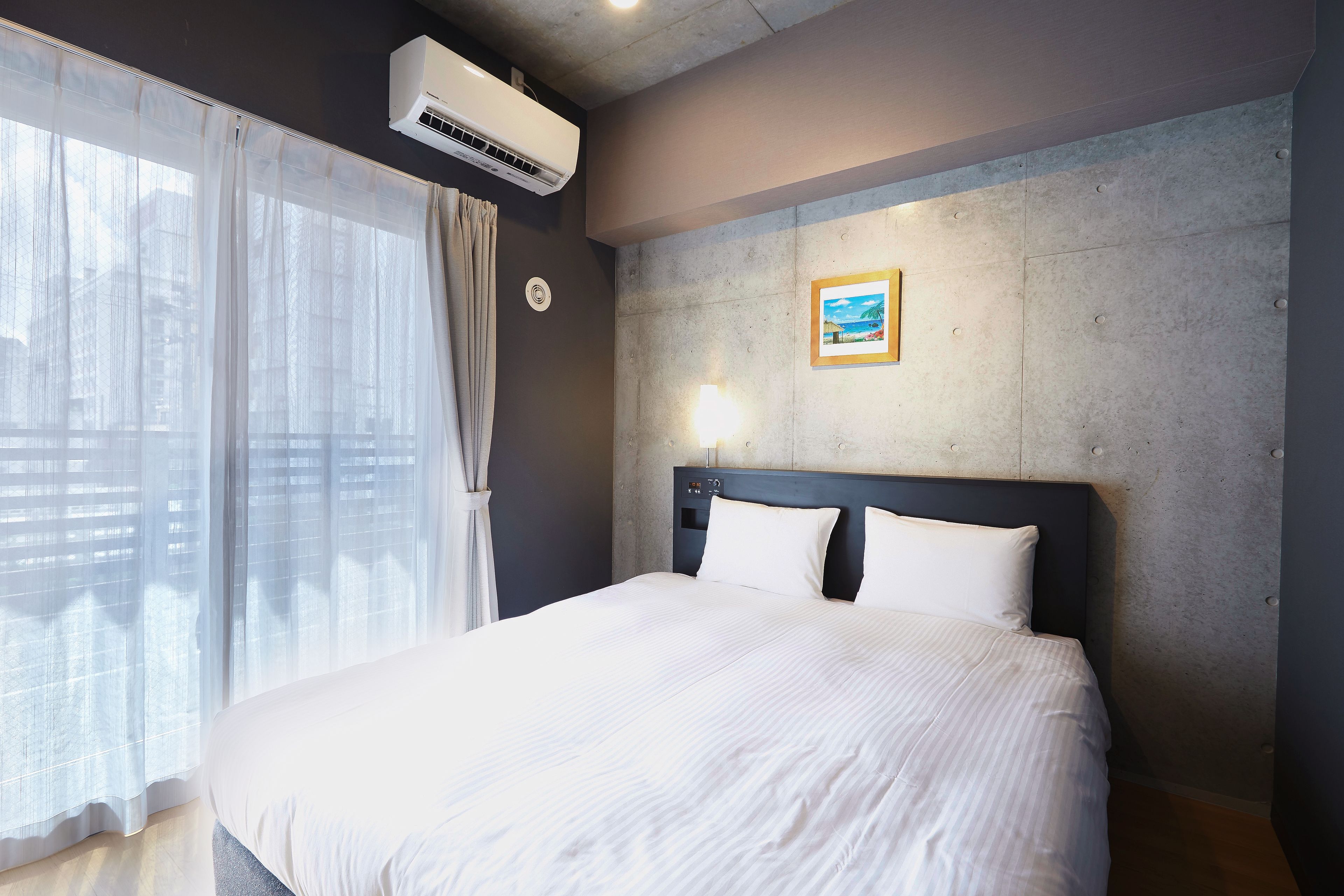 Habitación de hotel con una cama grande, una ventana con cortinas translúcidas, un equipo de aire acondicionado, una lámpara de pared y un cuadro enmarcado.