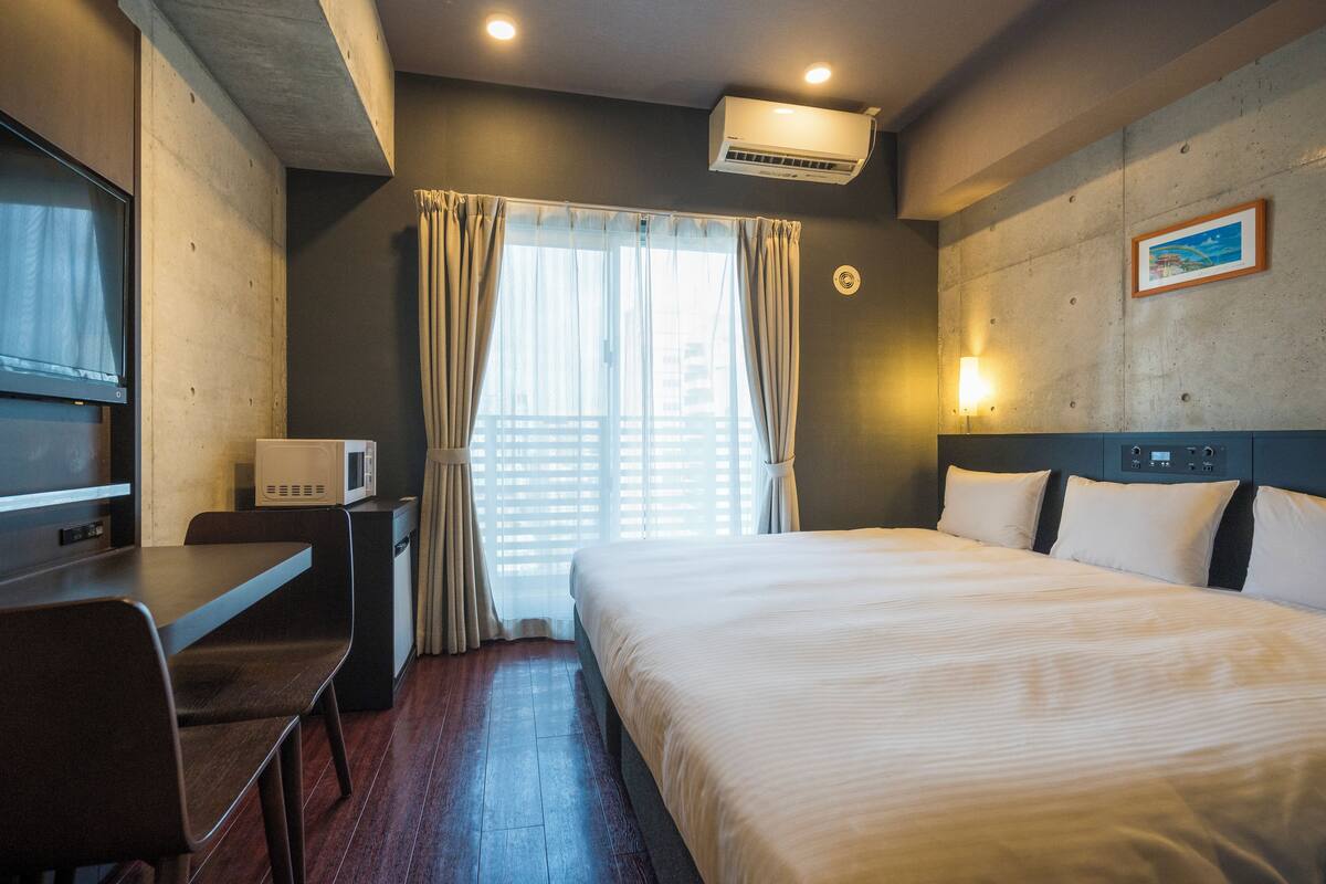 A hotel room with a bed, a desk with a chair, a microwave, a TV, a window with curtains, and an air conditioning unit.