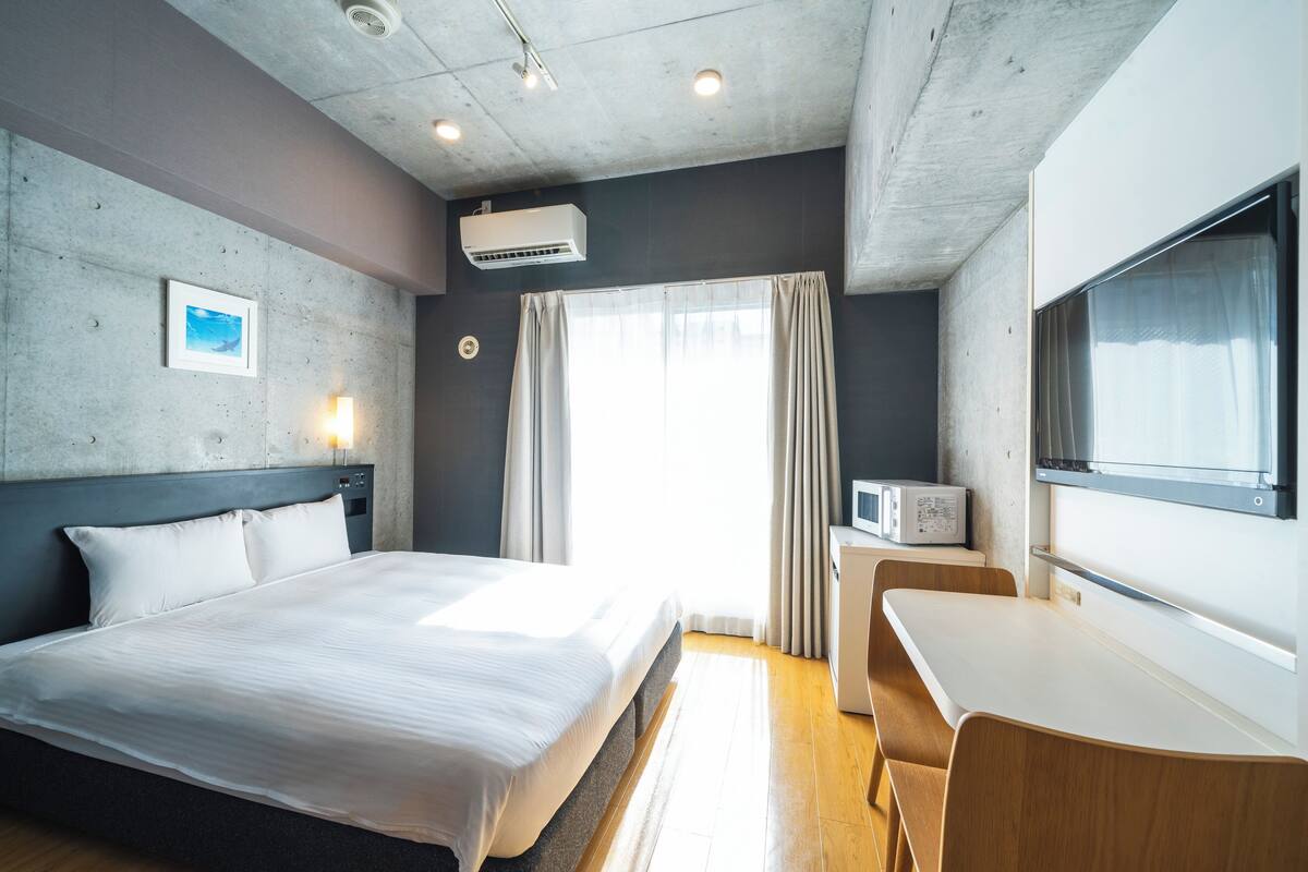 A modern hotel room with a large bed, a desk, a microwave, and a large window with curtains.