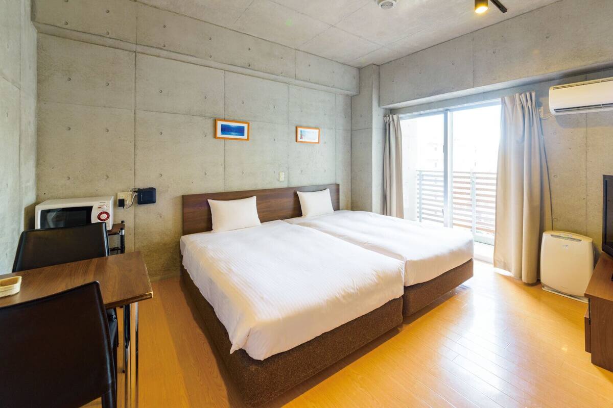 A hotel room with a double bed, a desk with a chair, a television, and an air conditioning unit.