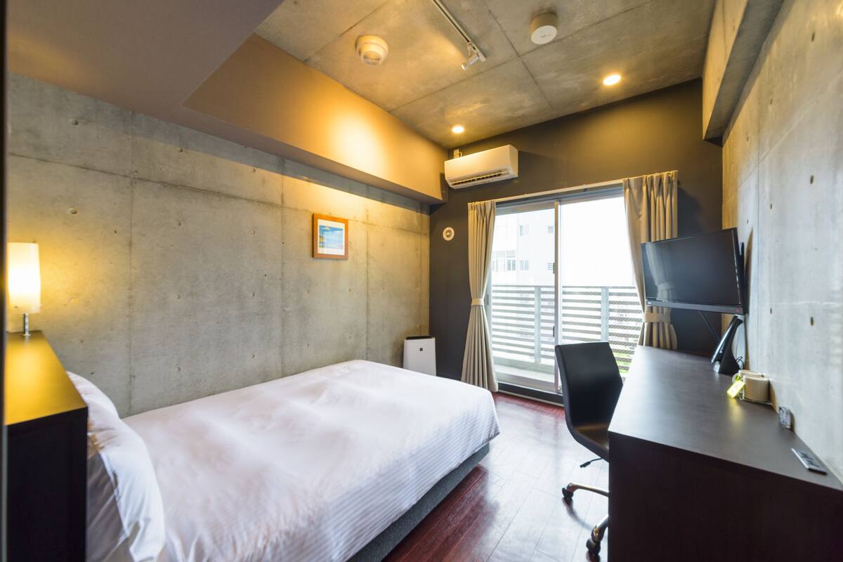 A hotel room with a bed, a desk with a chair, an air conditioning unit, and a window with curtains.