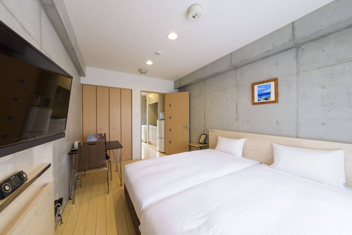 A hotel room with a bed, a desk, a chair, a small table, and a picture on the wall.