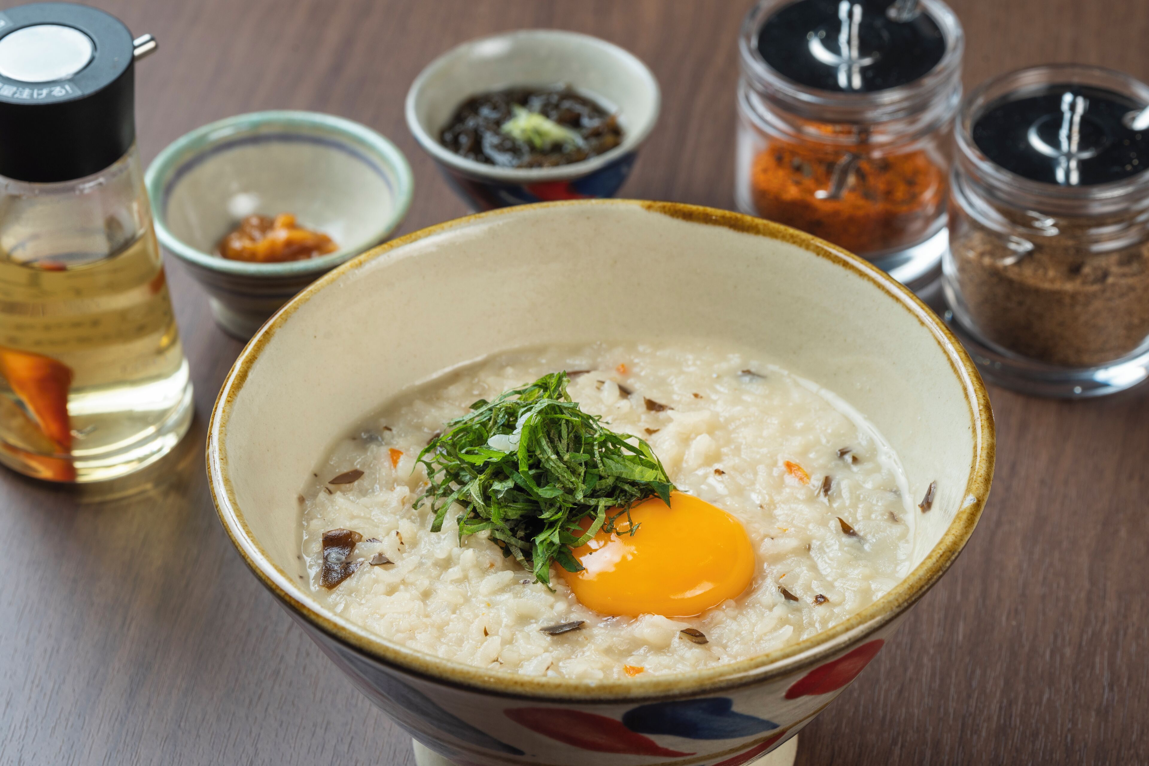 A bowl of porridge with an egg and greens, surrounded by condiments.