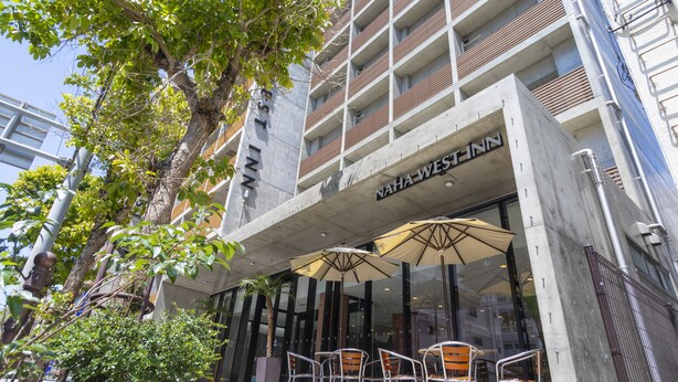 Un edificio moderno con un cartel que dice "NAHA WEST-NN". Hay sillas y sombrillas en el exterior, frente a la entrada.