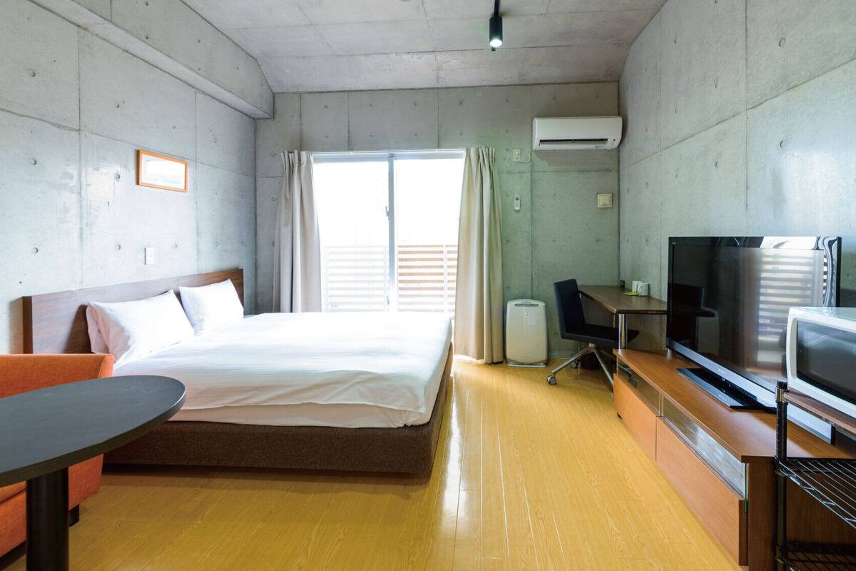 A hotel room with a bed, a desk, a TV, and an air conditioning unit.