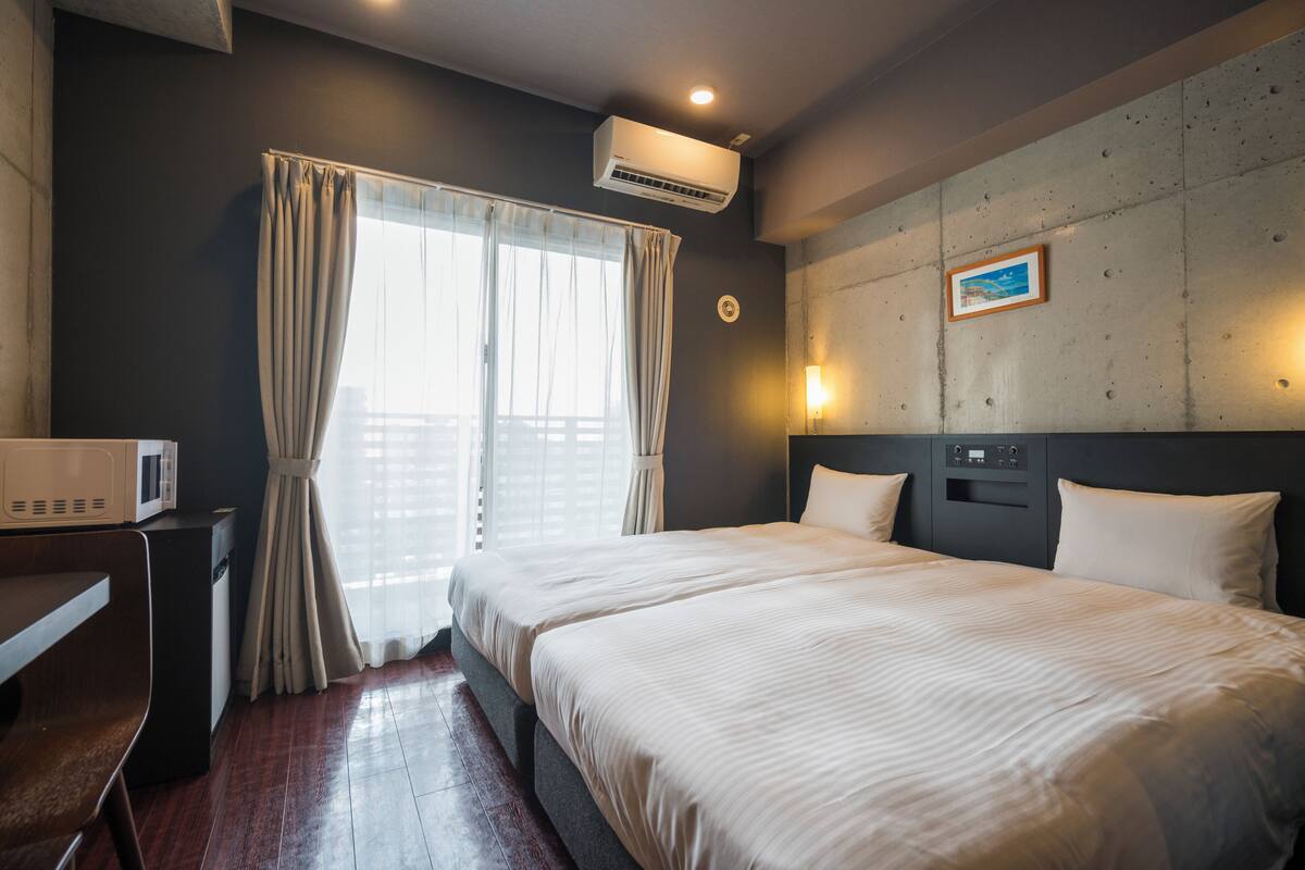 A hotel room with a bed, a desk, an air conditioner, a window with curtains, and a wall-mounted light fixture.