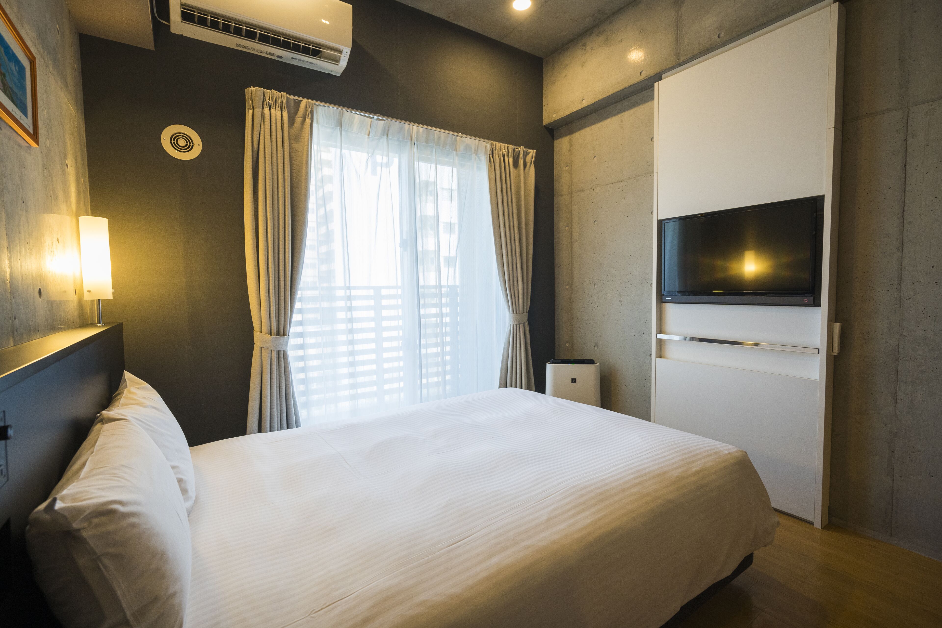 Habitación de hotel con cama, televisor montado en la pared, ventana con cortinas y aire acondicionado.