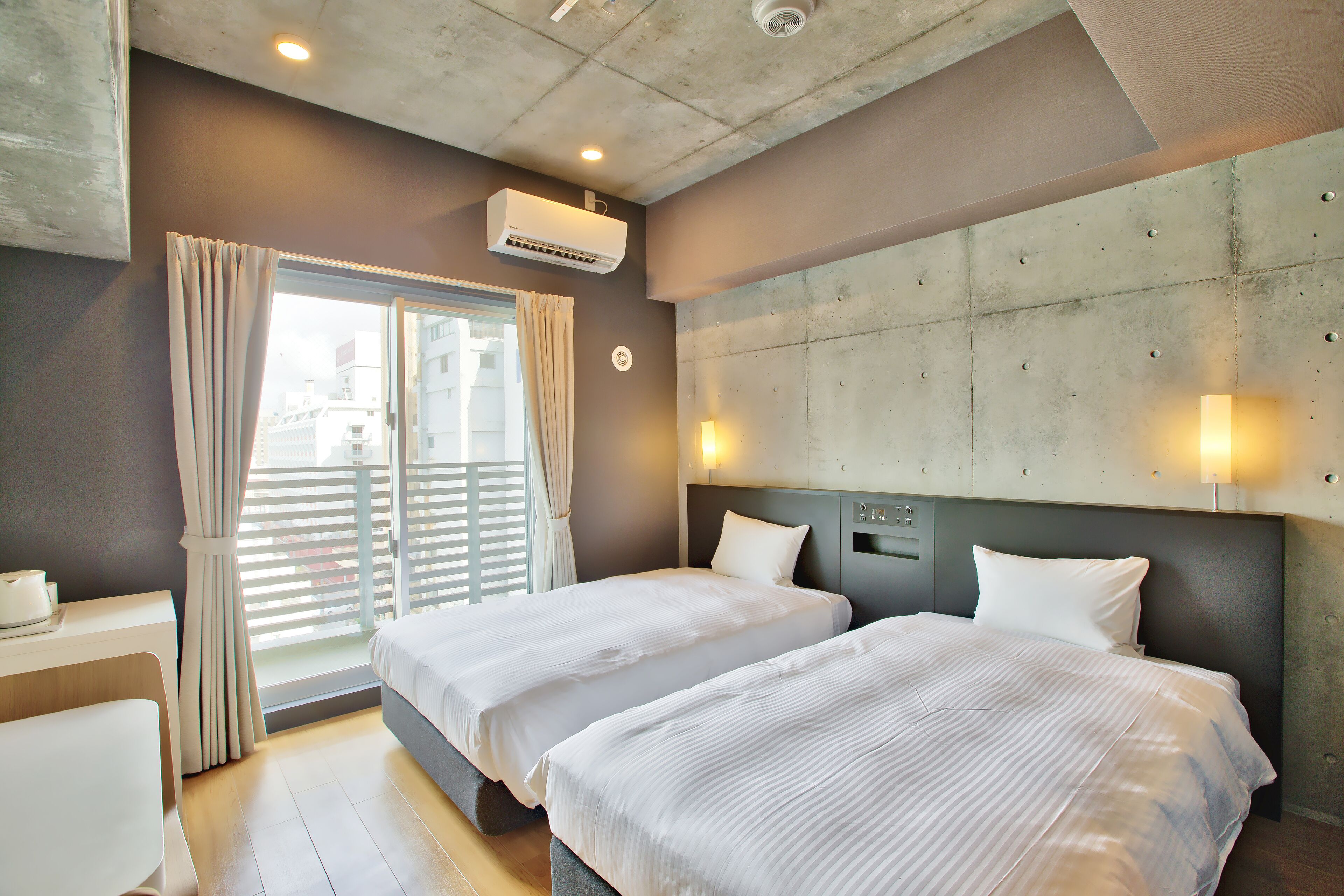 Hotelkamer met twee bedden, een raam met gordijnen, een airconditioning en een betonnen accentmuur met wandlampen.