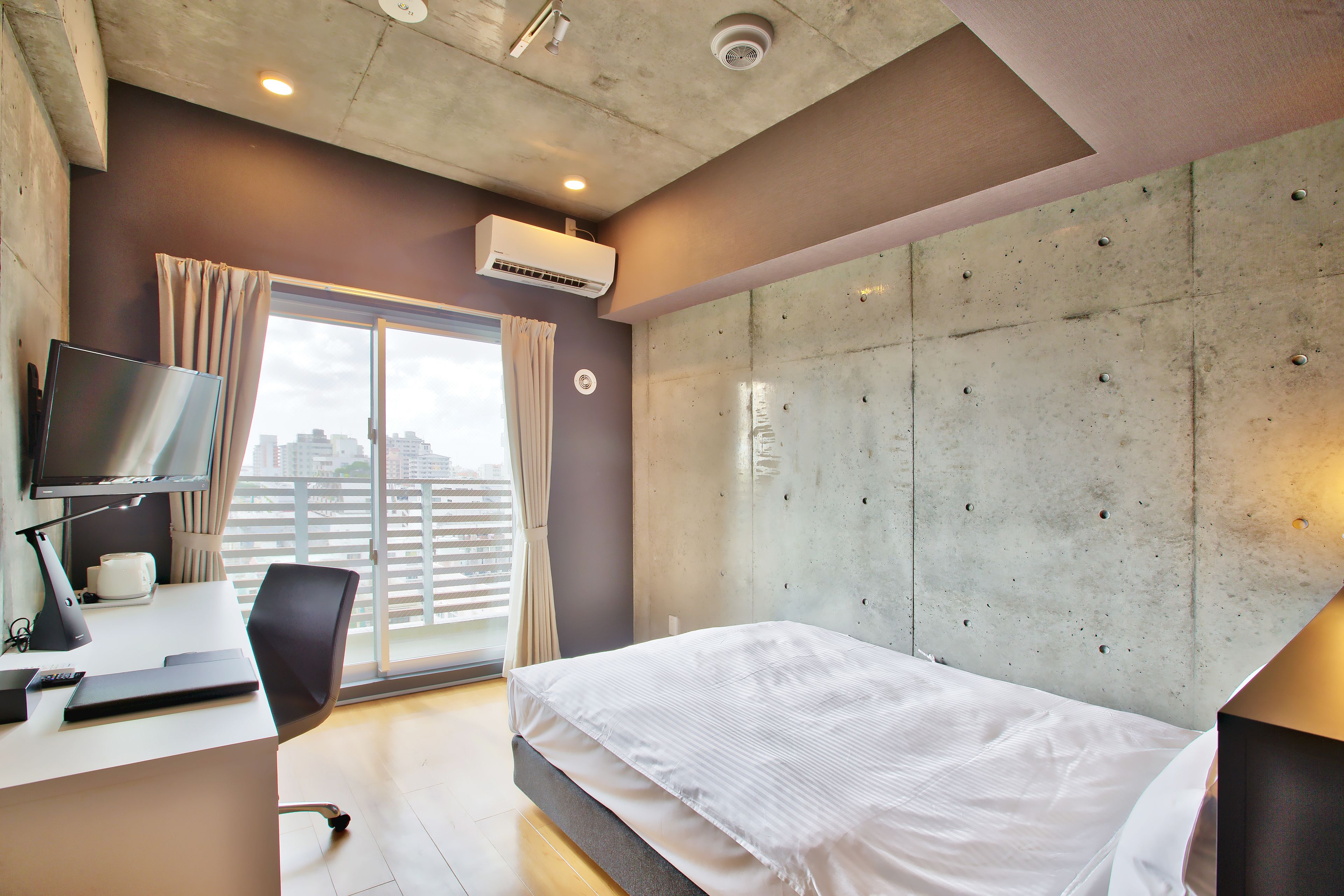 Ein modernes Hotelzimmer mit einem großen Bett, einem Schreibtisch mit Computer und Blick auf die Stadt durch das Fenster.