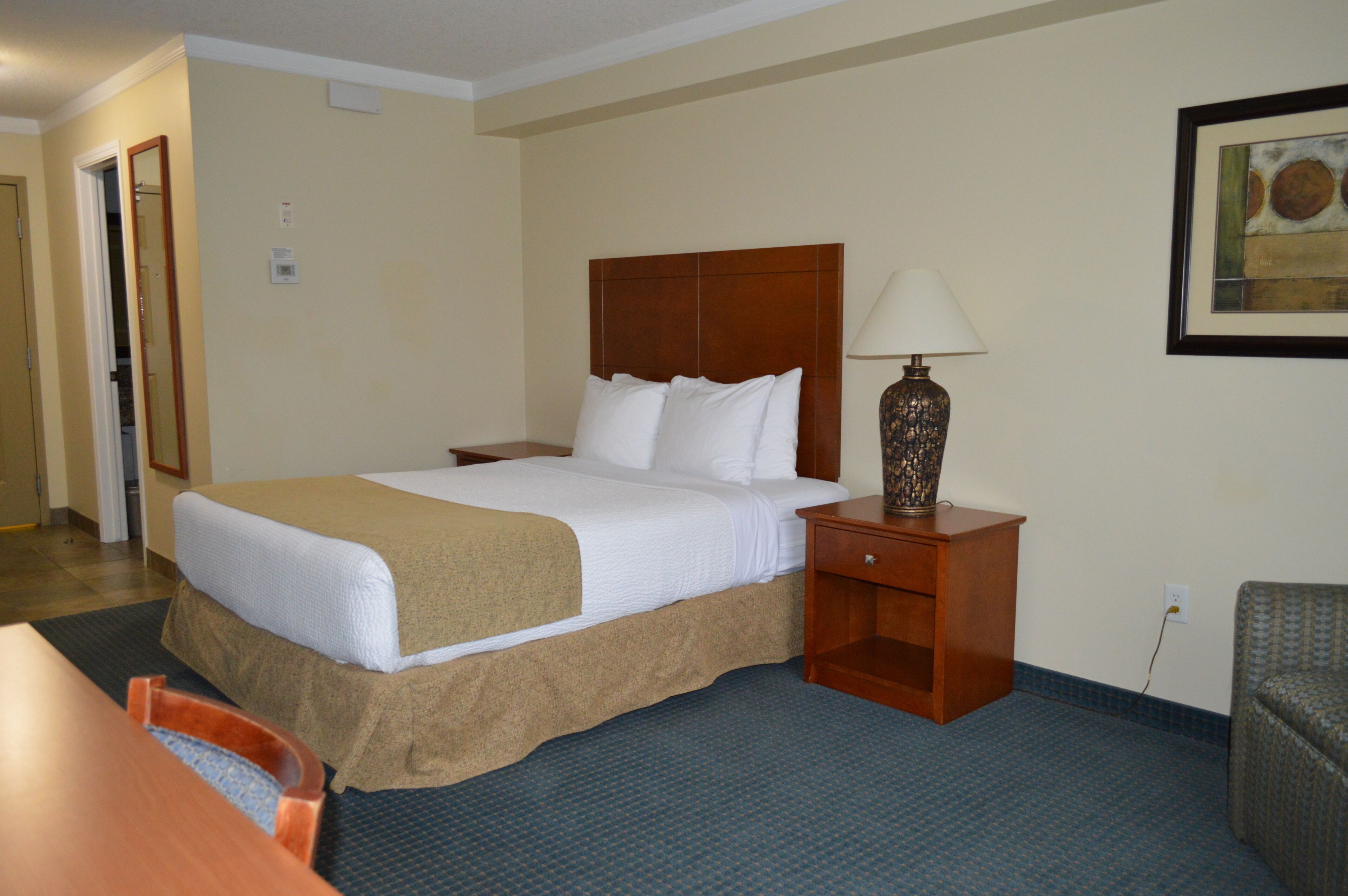 Habitación de hotel con una cama grande, una mesita de noche de madera con una lámpara, una silla y una pequeña mesa.