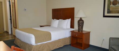 Habitación de hotel con una cama grande, una mesita de noche de madera con una lámpara, una silla y una pequeña mesa.