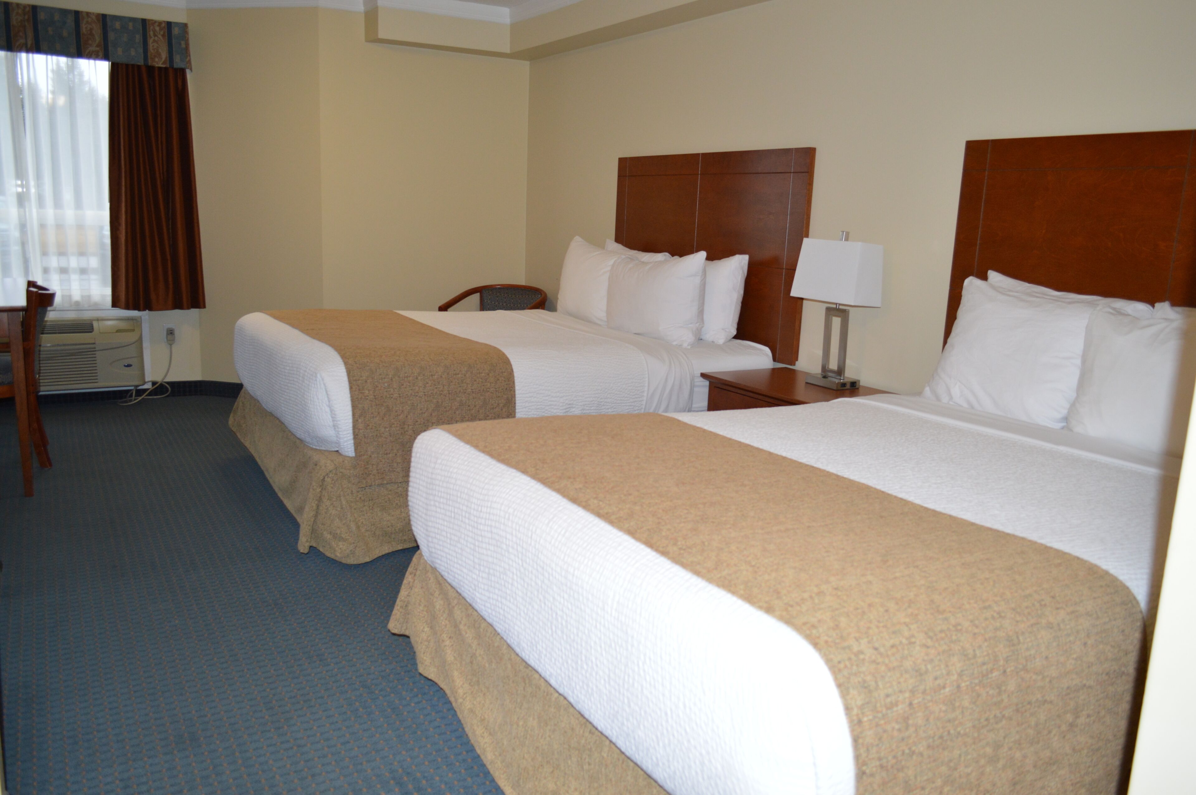 A hotel room with two beds, a desk, a chair, and a lamp.