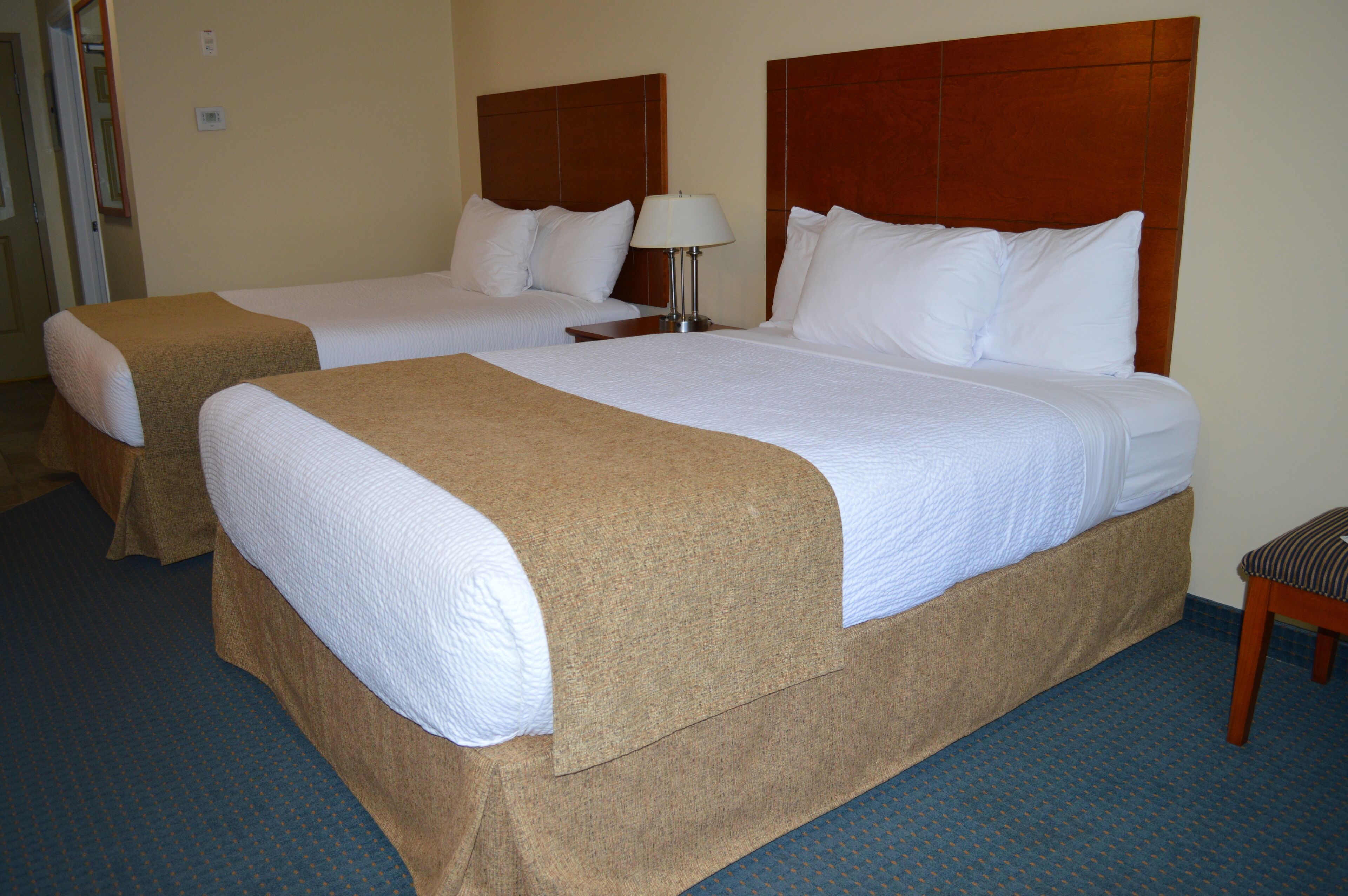 A hotel room with two beds, each with a wooden headboard, a bedside table with a lamp, and a bench by the window.