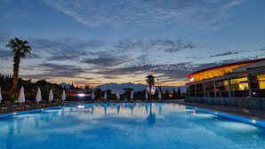 Una piscina con tumbonas y sombrillas, un edificio con techo curvo, y montañas al fondo.