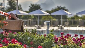 Ett område vid poolen med en solstol under ett parasoll med texten ”Nashira City Resort Hotel”, omgivet av frodig grönska och färgstarka blommor.
