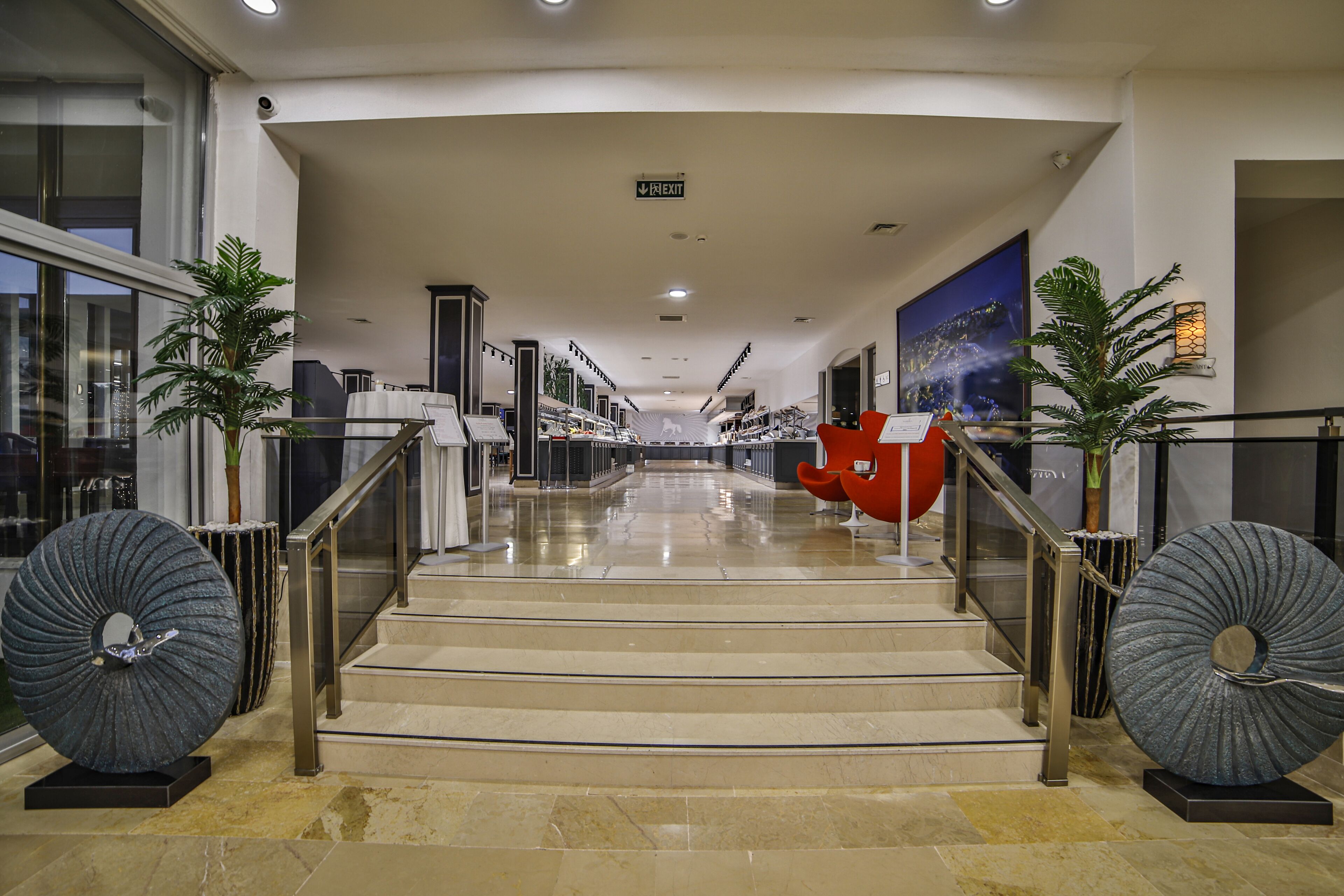 En rymlig och väl upplyst interiör med en central trappa, glasräcken och dekorativa element som stora skulpturer och krukväxter.