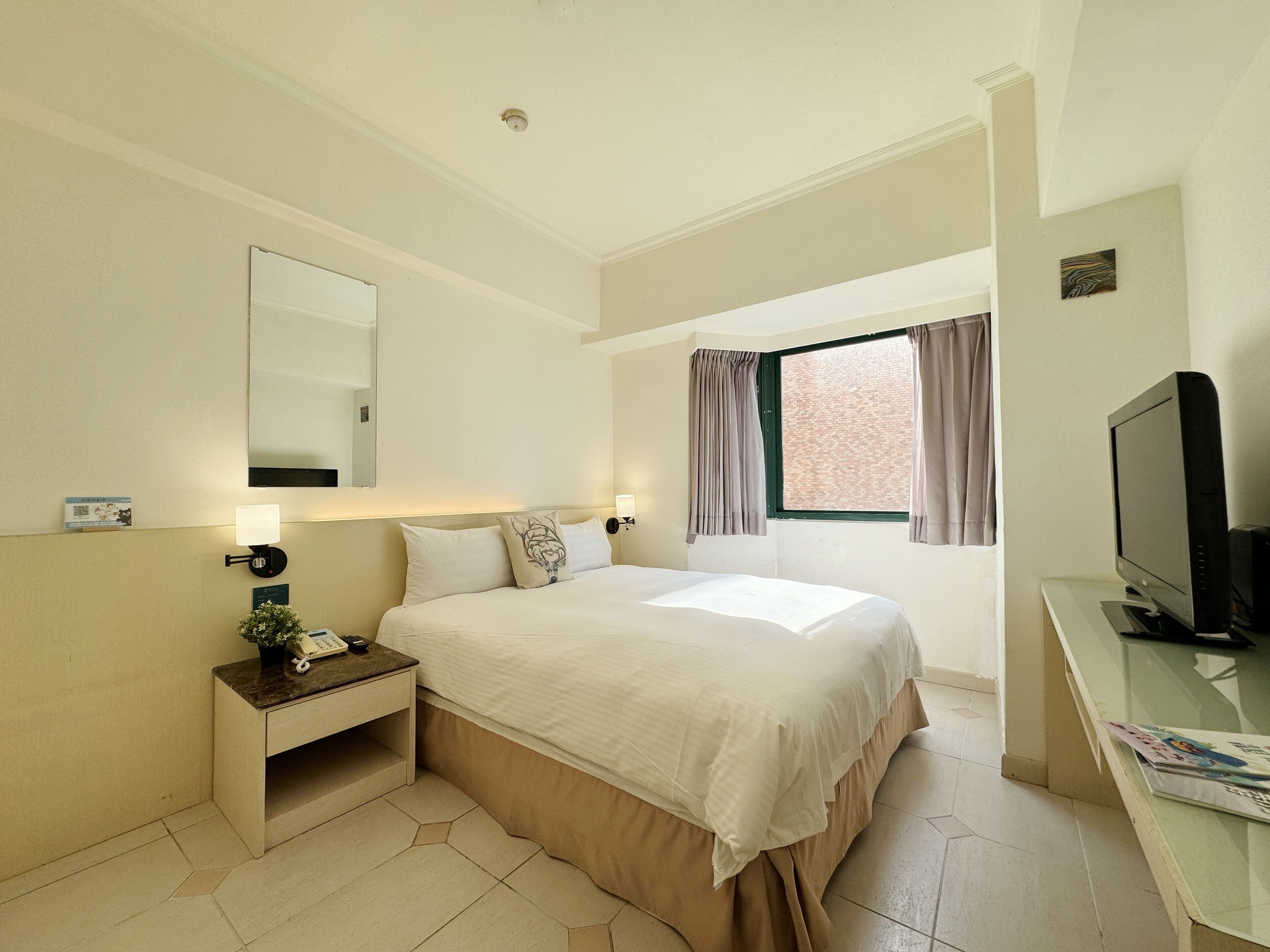 A hotel room with a bed, bedside table, lamp, TV, and a window with curtains.