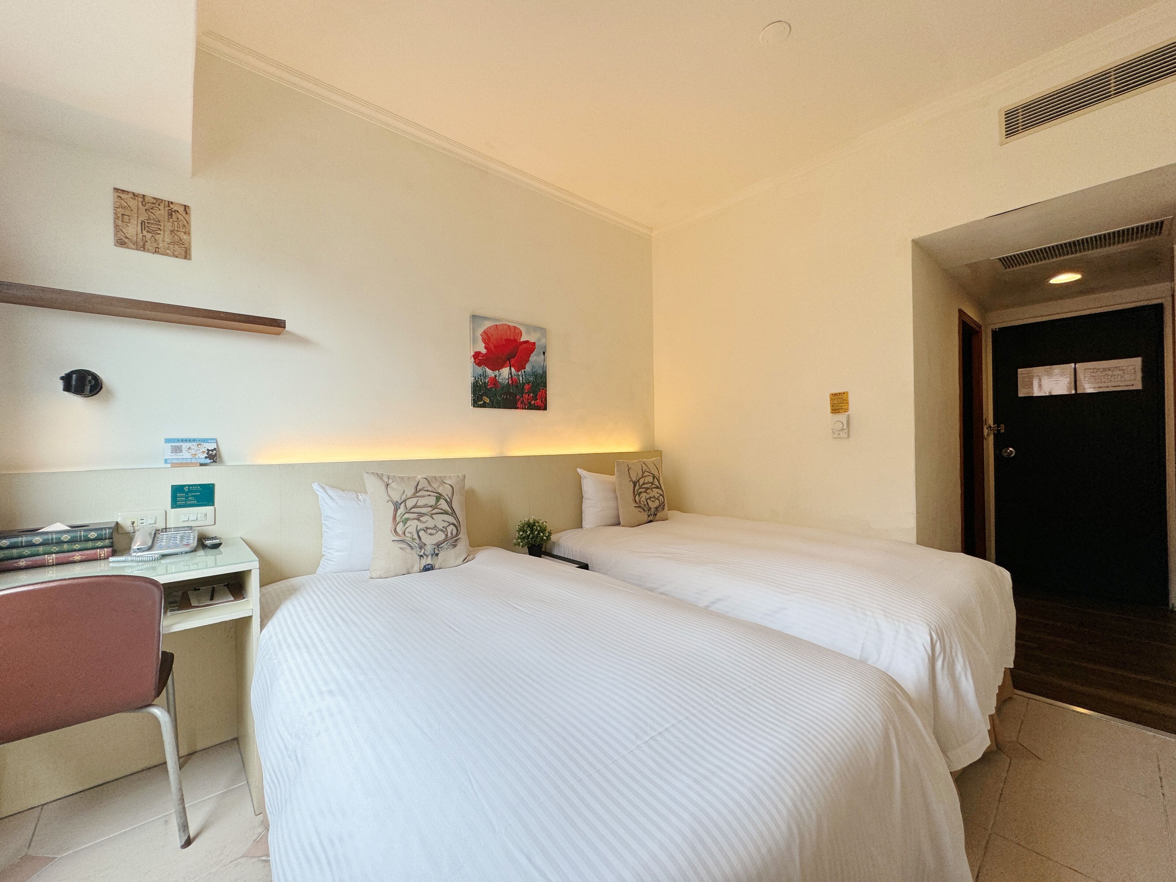 A hotel room with two beds, a desk, and a chair. There is a painting on the wall and a door with a sign.
