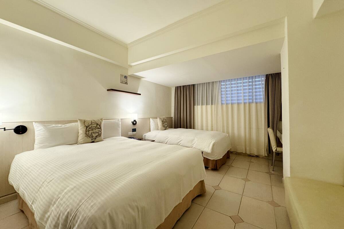 Una habitación de hotel con dos camas, un ventanal grande con cortinas y suelo de baldosas.