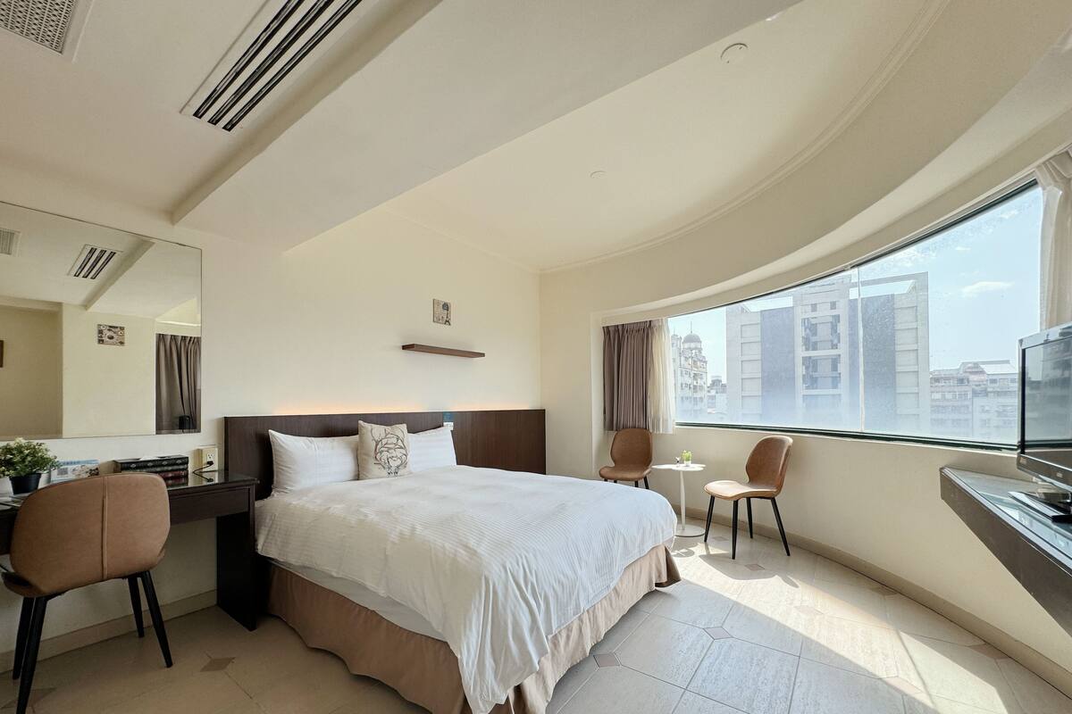 Habitación de hotel con una cama grande, un escritorio con silla, una mesita y un ventanal con vistas a la ciudad.