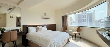 Habitación de hotel con una cama grande, un escritorio con silla, una mesita y un ventanal con vistas a la ciudad.