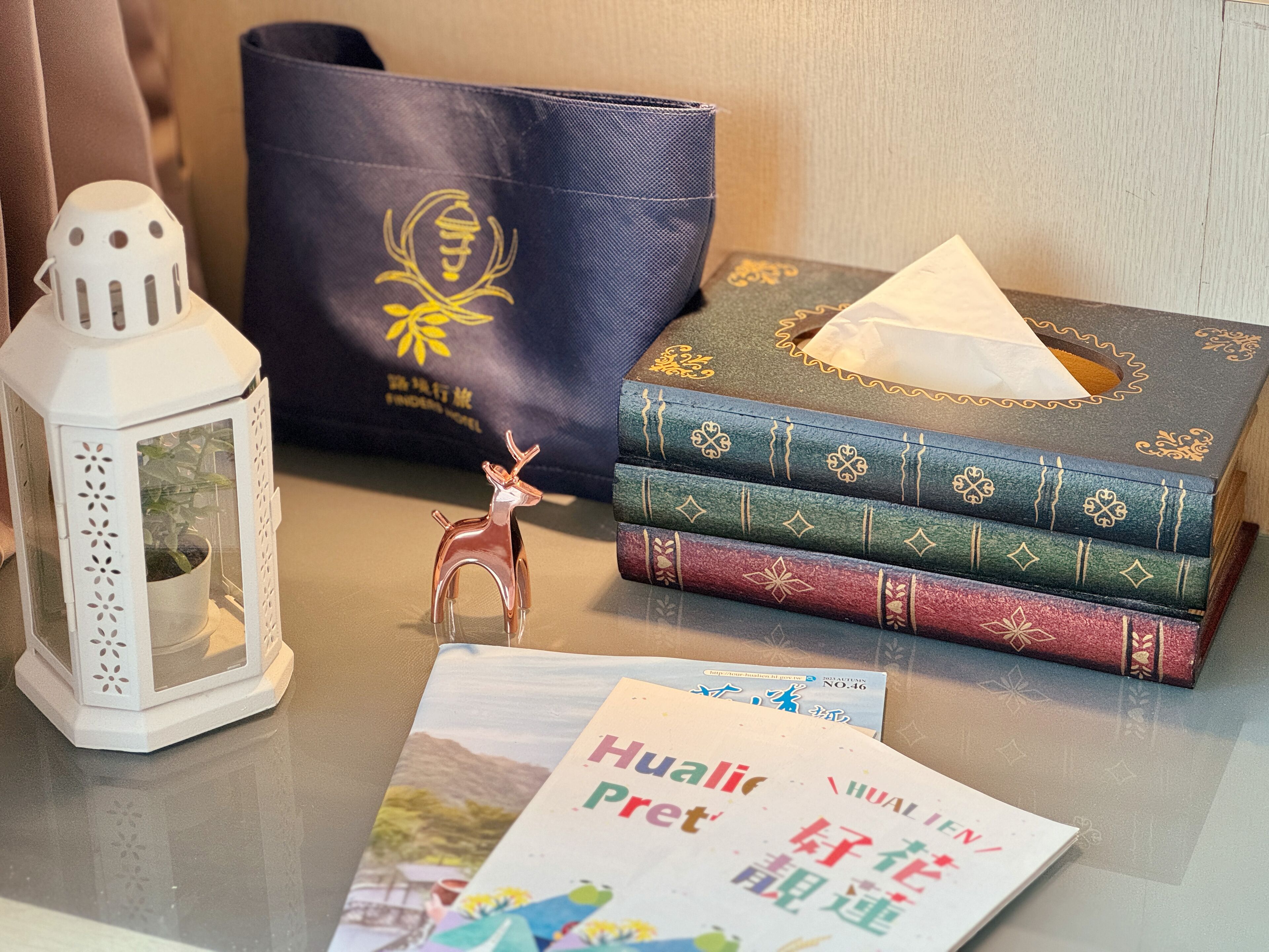 Una mesa con un farol blanco, una figurita decorativa de ciervo, una pila de libros, un caja de pañuelos y un folleto.