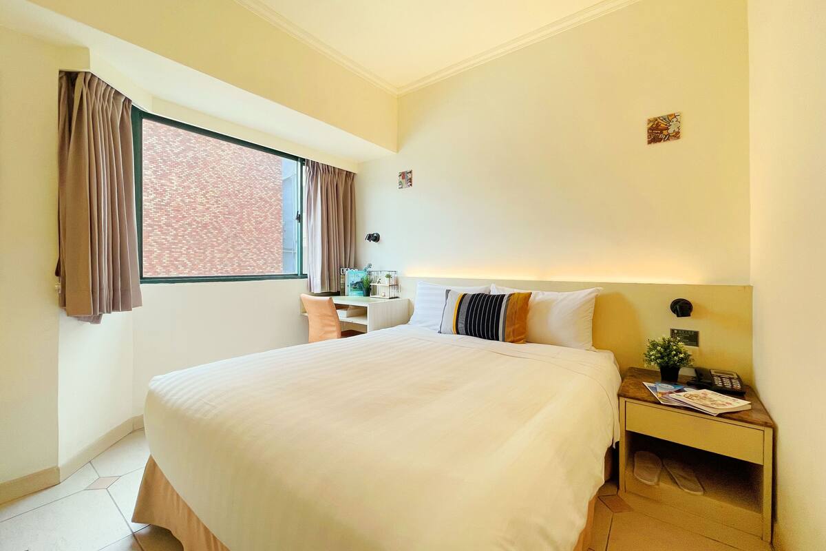 Habitación de hotel con cama, escritorio, mesita de noche, ventana con cortinas y una lámpara en la pared.