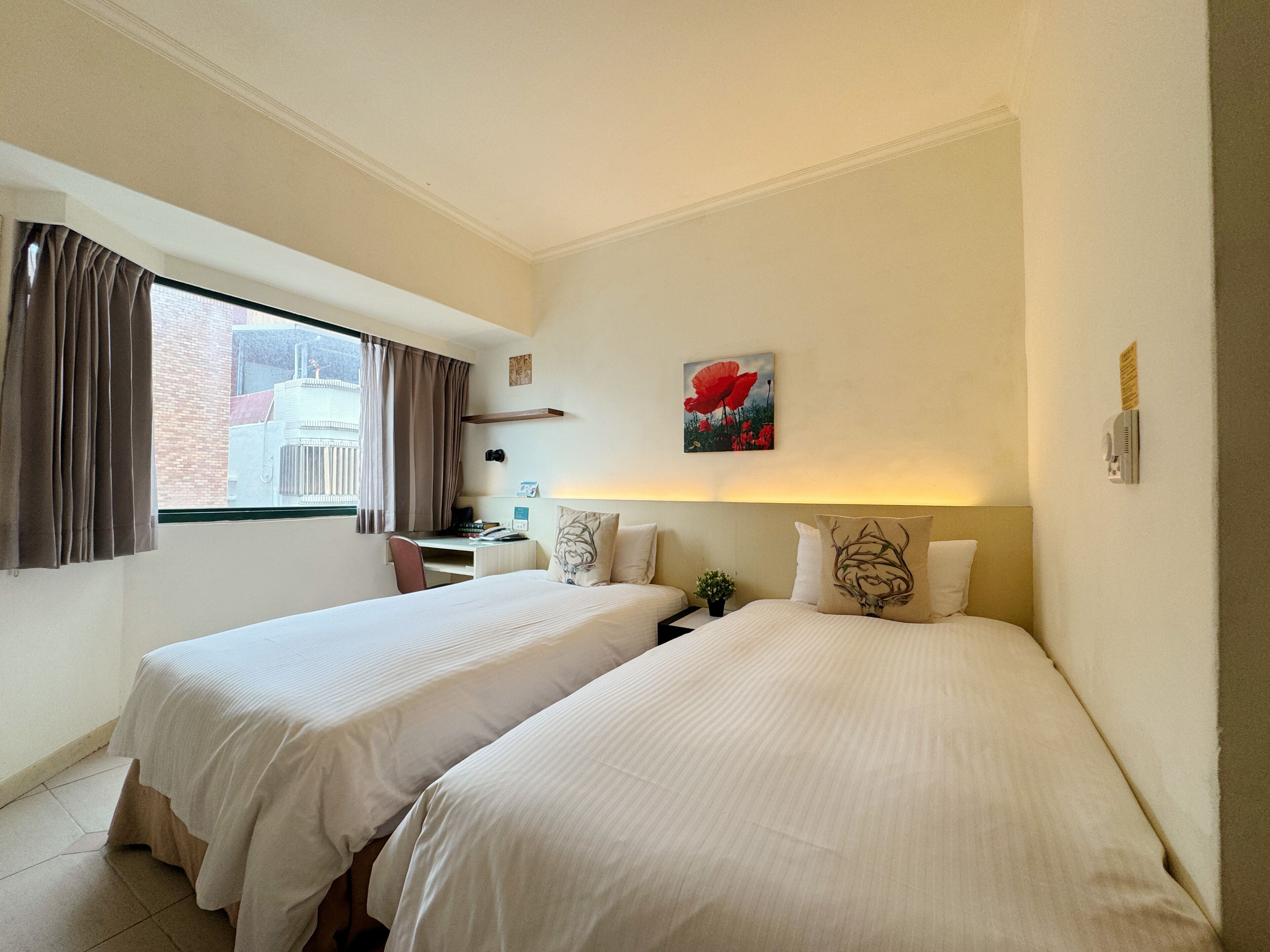 A hotel room with two beds, a desk, a chair, a window with curtains, a painting, and a small plant.