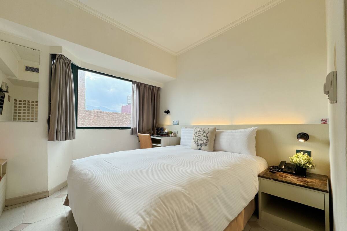 Una habitación de hotel con una cama grande, un escritorio con silla, una mesita de noche con lámpara y vistas a la ciudad a través de la ventana.