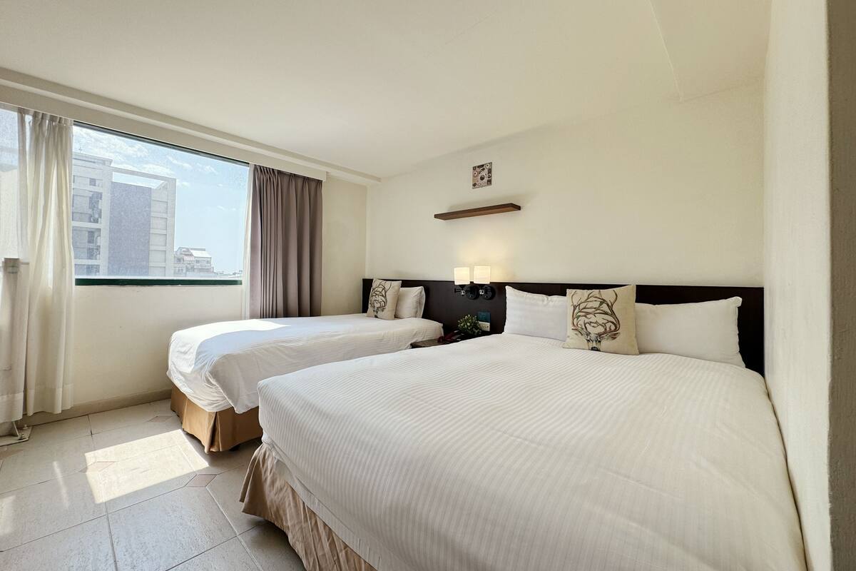 Habitación de hotel con dos camas, una ventana con cortinas, una mesita de noche con lámpara y un estante en la pared.