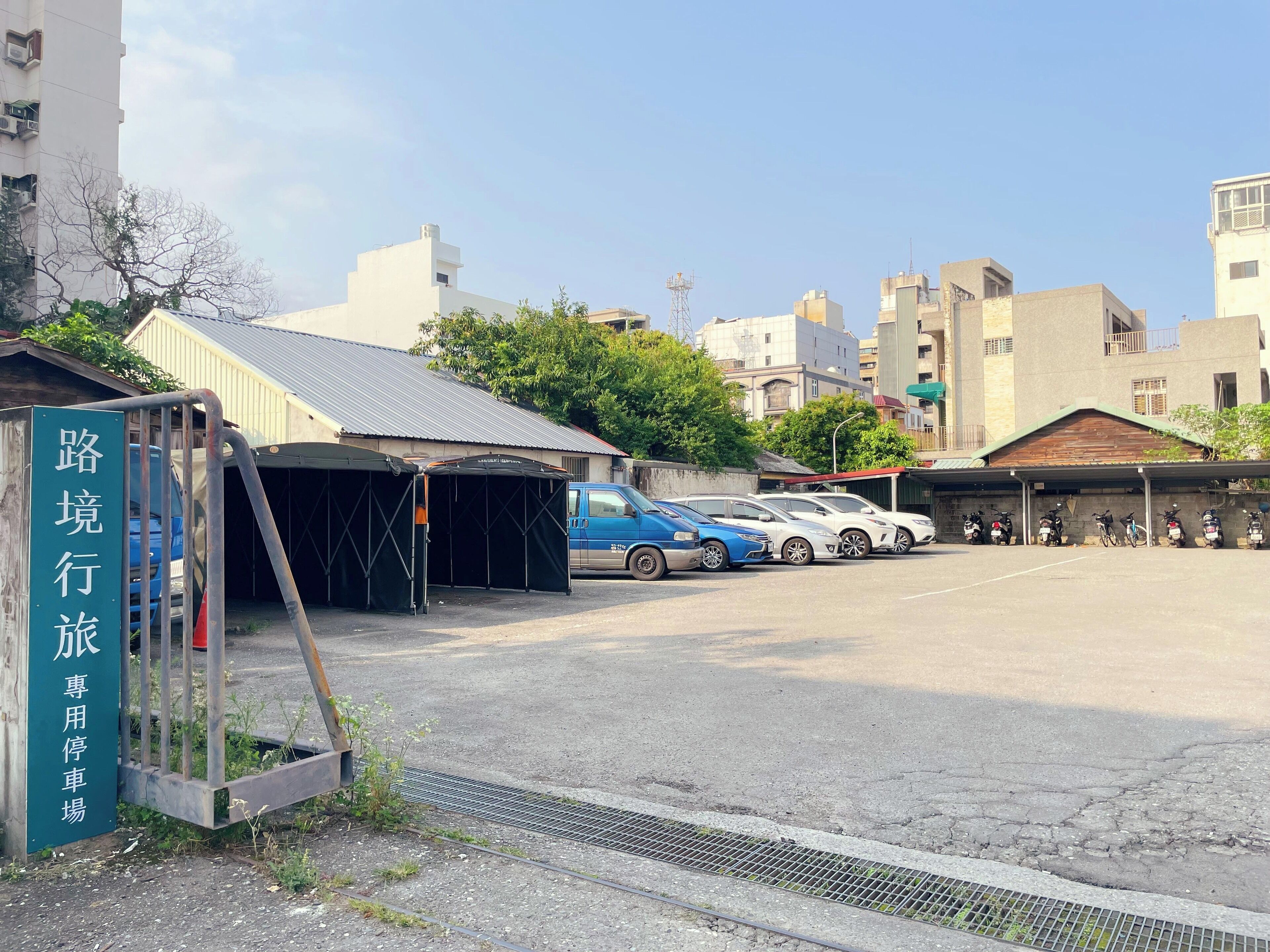 A parking area with a green sign, a blue van, and several cars and motorcycles parked.
