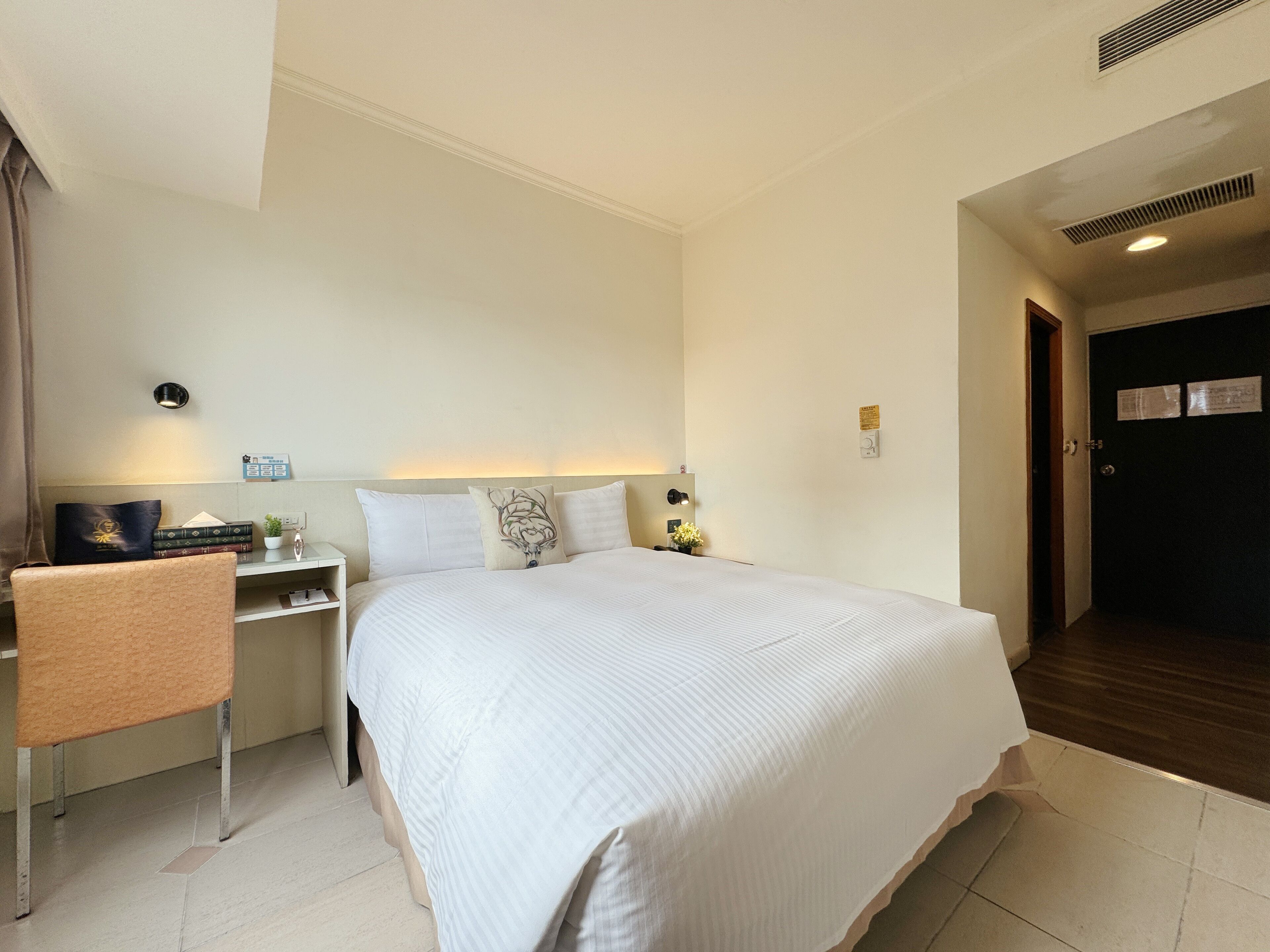 A hotel room with a large bed, a desk with a chair, a nightstand, and a wall-mounted light.